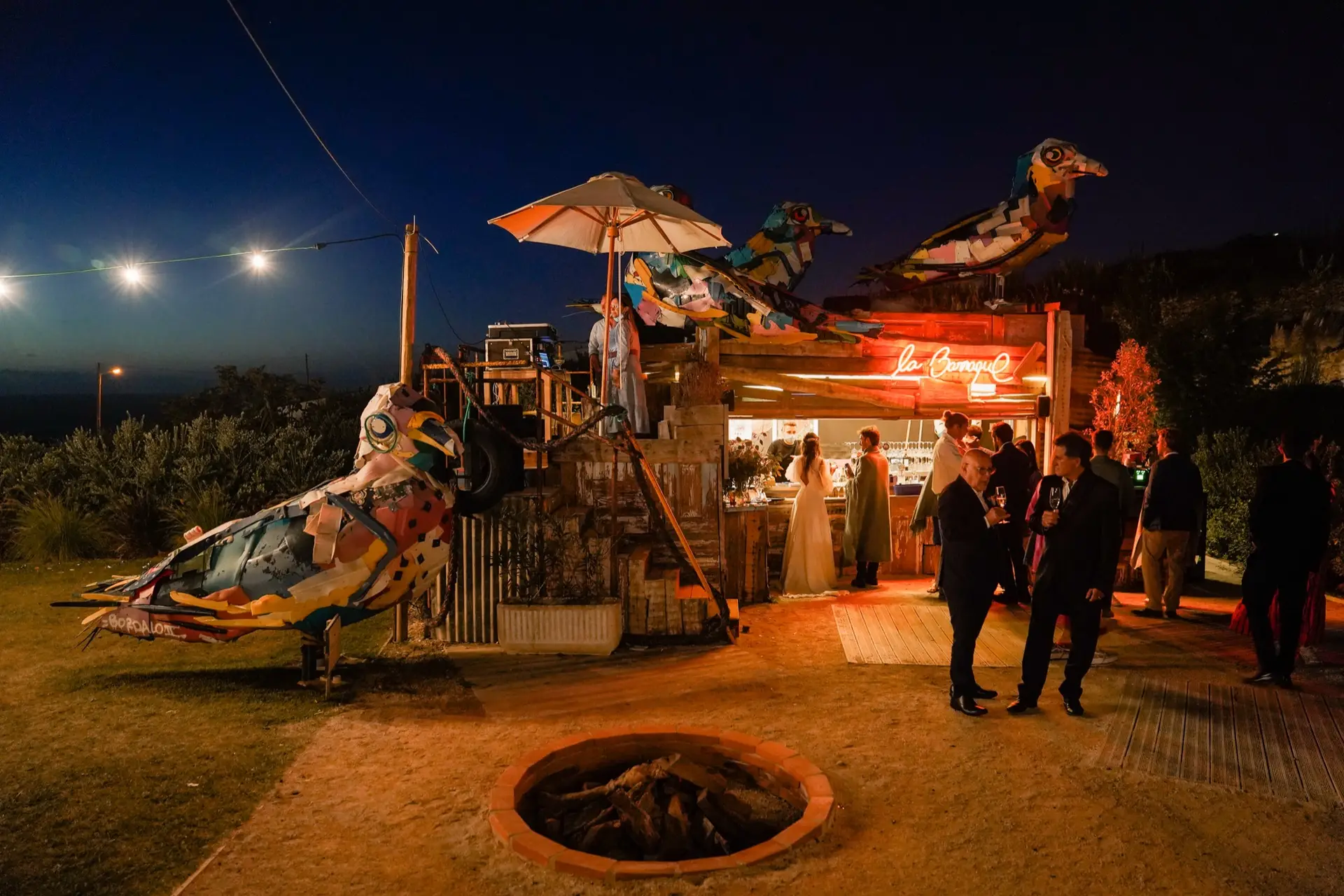 O bar La Barraque à noite, com os pássaros coloridos, luzes suspensas e convidados à volta da fogueira